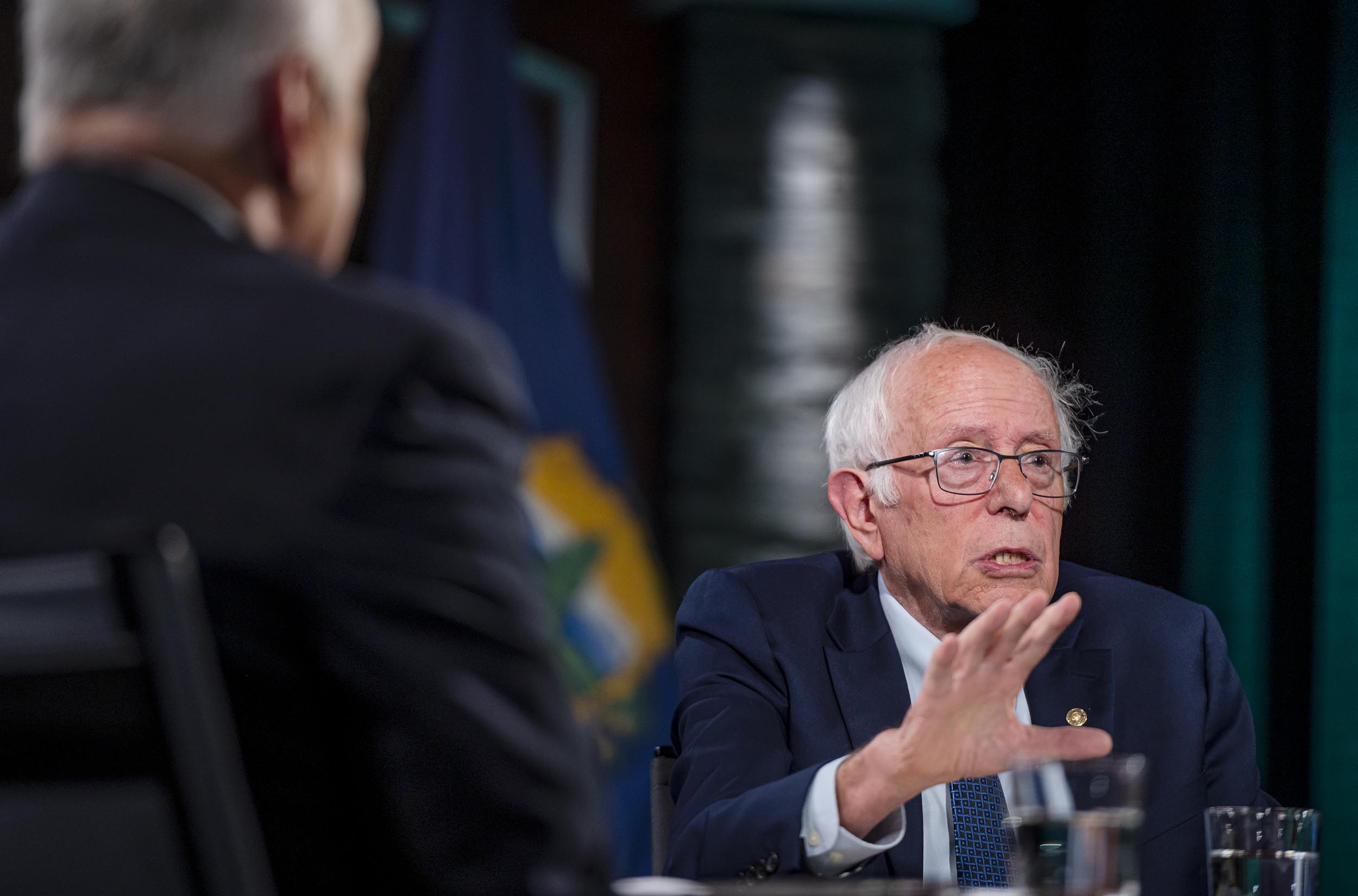 An elderly man in glasses and a suit gestures while speaking at a table. Another person is partially visible in the foreground.
