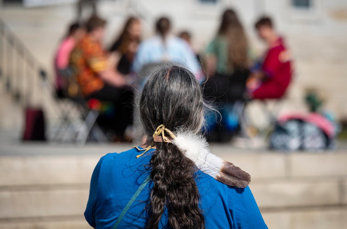 A person with a braid and a feather in their hair watches a group of people sitting in a circle on a set of steps.