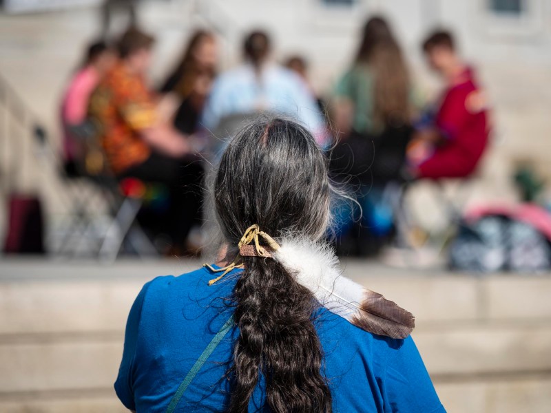 A person with a braid and a feather in their hair watches a group of people sitting in a circle on a set of steps.