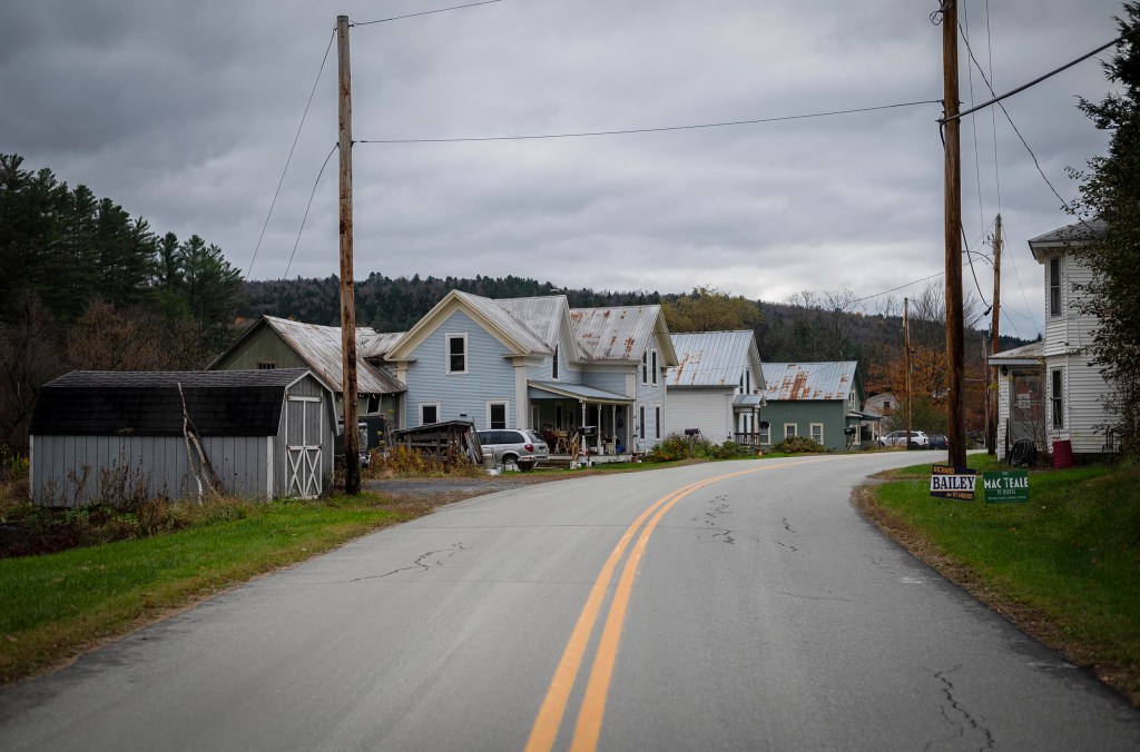 A quiet, rural street curves past a line of weathered houses with tin roofs under an overcast sky. Campaign signs are visible on the roadside.