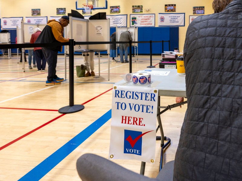 People voting in a gymnasium, with a "Register to Vote Here" sign on a table in the foreground.