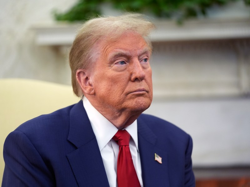 An older man in a suit and red tie sits in a room with a white fireplace and green foliage in the background.