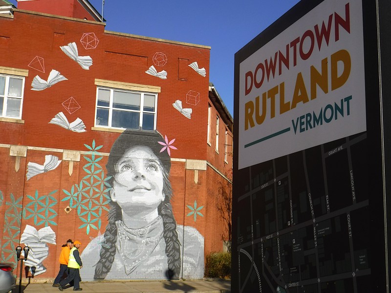 Mural of a girl with braided hair looking up, surrounded by books and geometric shapes, on a red brick wall. Nearby sign reads "Downtown Rutland Vermont." Two people walk on the sidewalk.