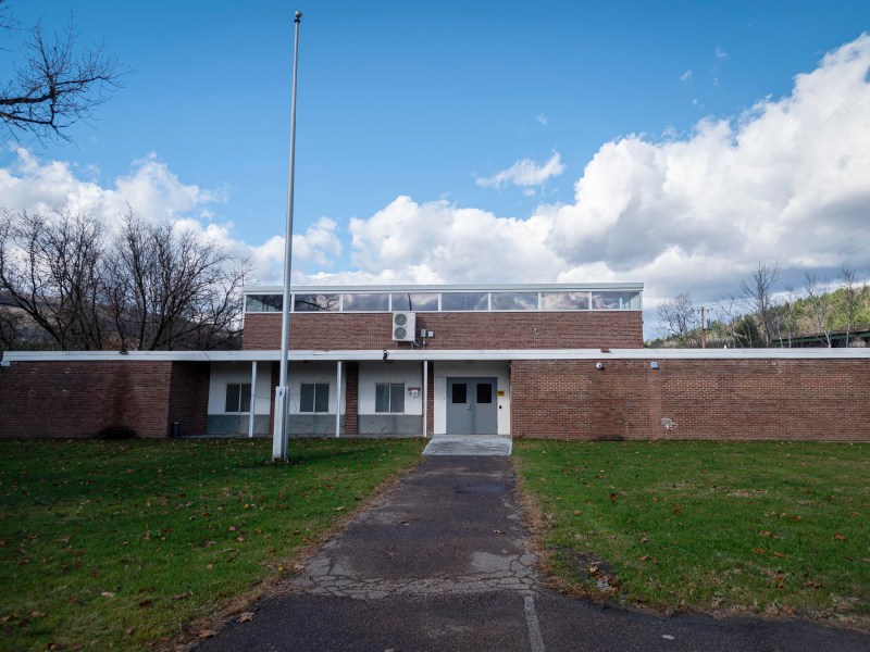 A two-story brick building with a flat roof and a flagpole in front, surrounded by grass and trees under a partly cloudy sky.
