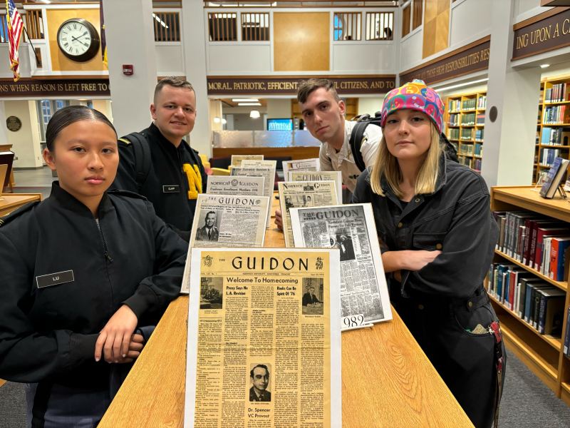 Four people stand in a library near a display of vintage newspapers titled "Guidon.