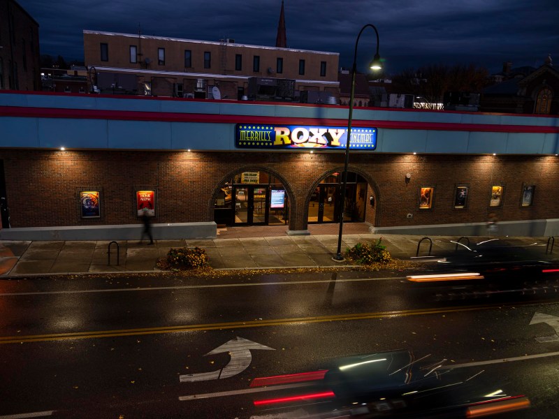 A movie theater at night with a lit marquee reading "Roxy," wet street in the foreground, and blurry car lights passing by.
