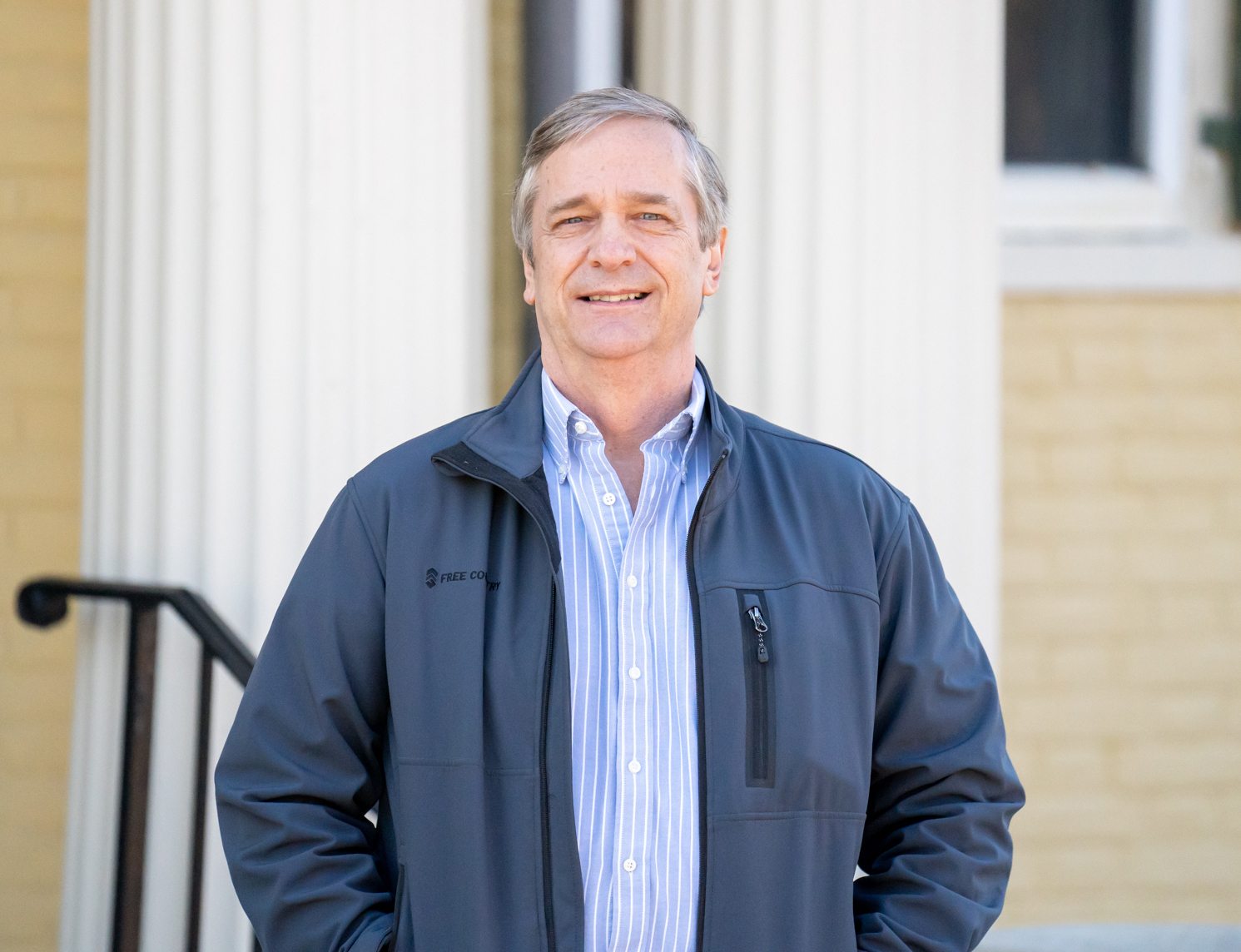 A man in a jacket stands in front of a building with columns, smiling at the camera.