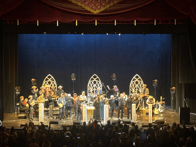 A live musical performance onstage with a band, various musicians, and singers. Stage lights illuminate performers and instruments against a decorative backdrop. Audience visible in the foreground.