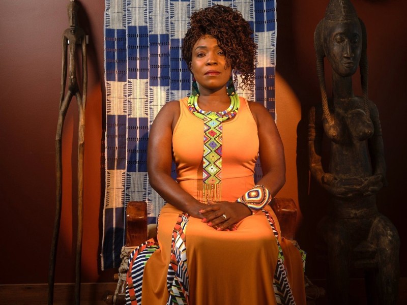 A woman in an orange dress sits on a chair, surrounded by African art and decor.