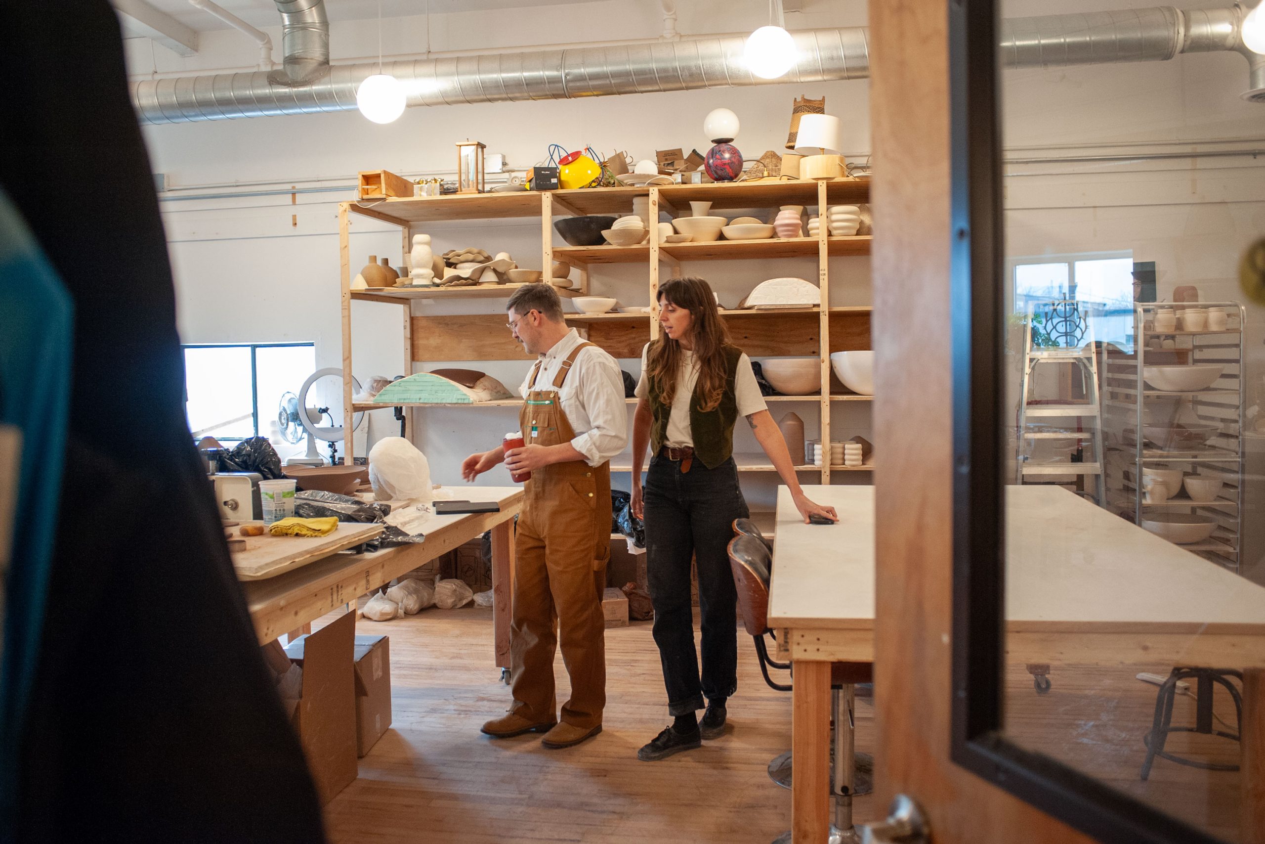 Two people stand in a workshop with shelves full of pottery and materials. They appear to be discussing something on the table in front of them.