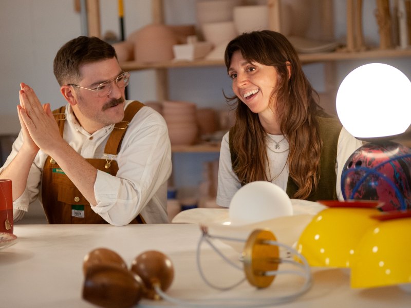 Two people sitting at a table with various colorful objects and a glowing lamp, shelves in the background.