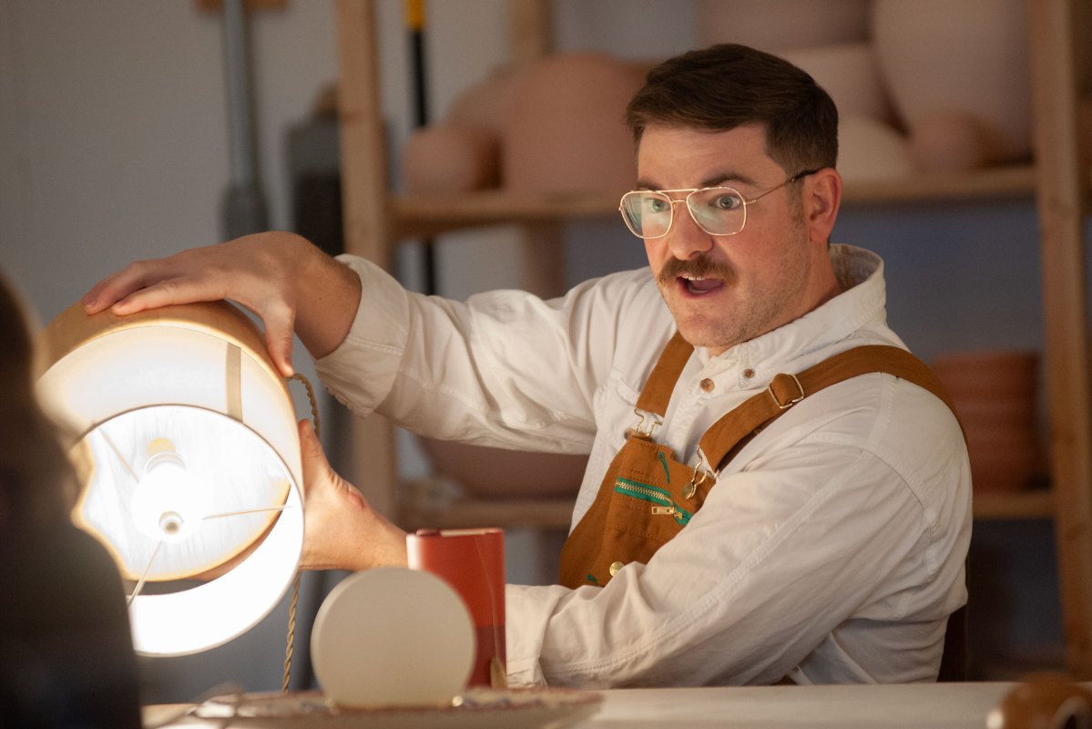 A man wearing glasses and overalls examines a lamp in a workshop.