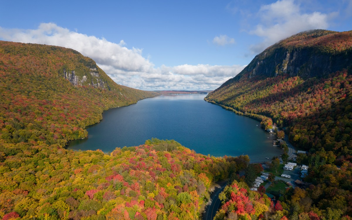 Aerial view of a lake surrounded by forested mountains in autumn, with vibrant red and orange foliage. A few buildings are visible near the shore.
