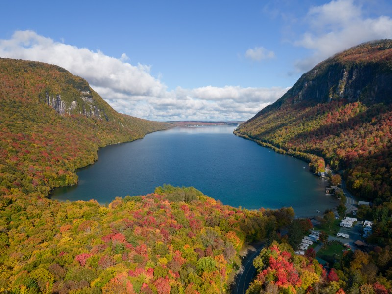 Aerial view of a lake surrounded by forested mountains in autumn, with vibrant red and orange foliage. A few buildings are visible near the shore.