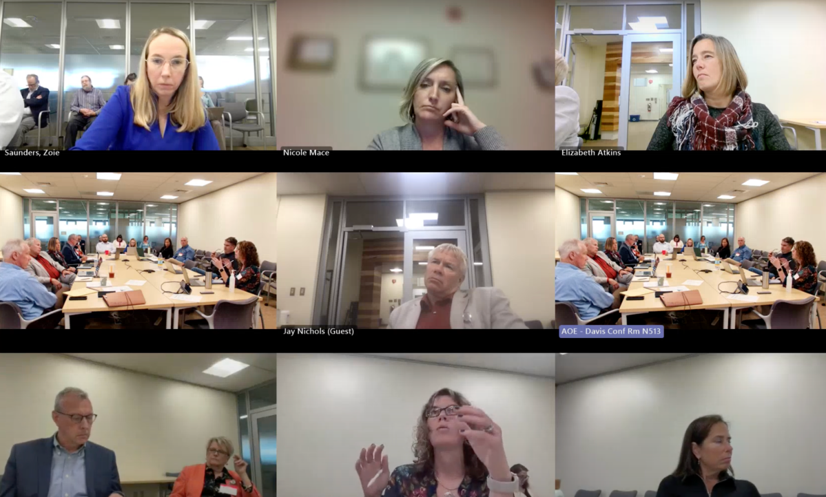 A video conference with nine participants in a meeting room setup. Some are seated at a table, while others appear on screens from different locations.