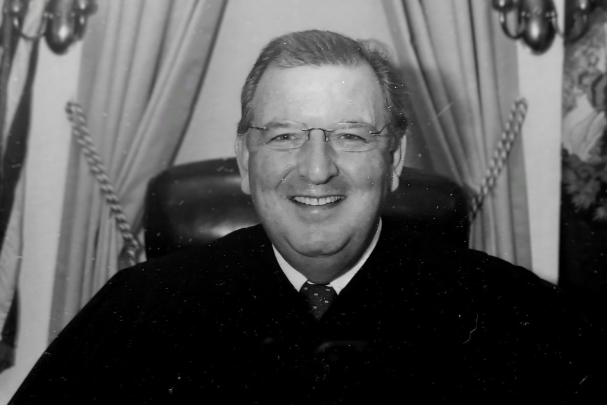 A man wearing glasses and a suit smiles while sitting in an office.