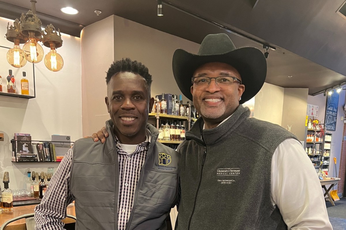 Two men smiling in a bar, one wearing a vest with a patch and the other in a cowboy hat and glasses. Shelves with bottles are in the background.