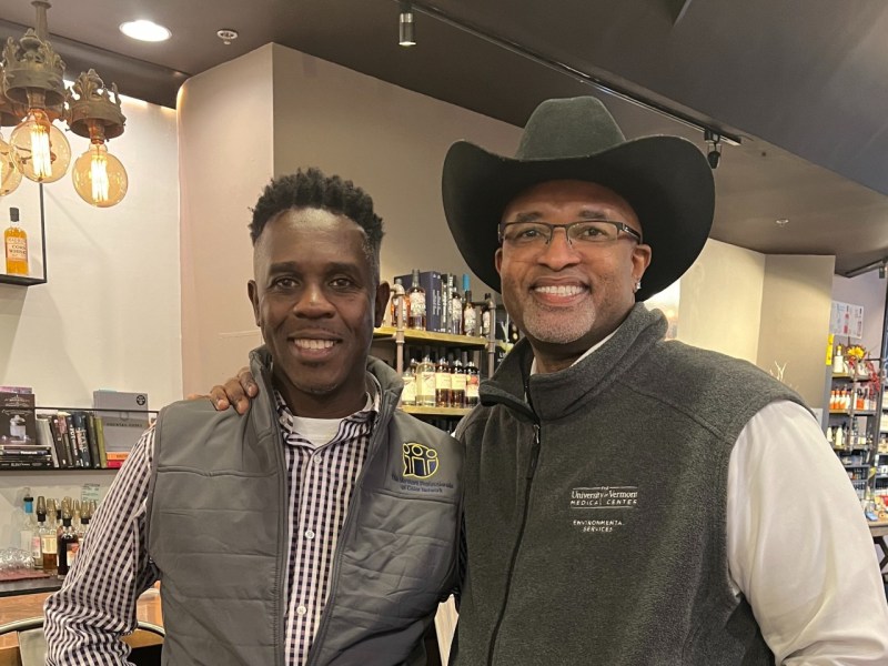 Two men smiling in a bar, one wearing a vest with a patch and the other in a cowboy hat and glasses. Shelves with bottles are in the background.