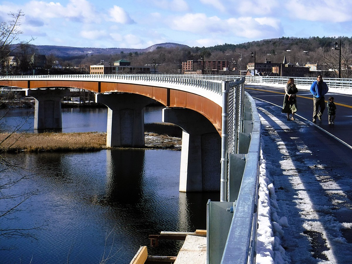A bridge crosses a river, with a person pushing a stroller and another walking a dog along a snow-dusted path. Buildings and hills are visible in the background.