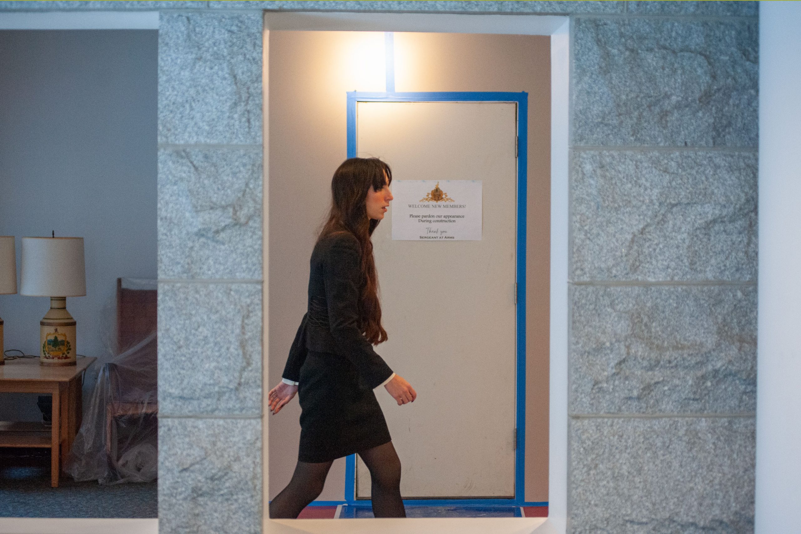 Person walking in a hallway, passing a taped-up door with a sign, stone walls visible on either side.