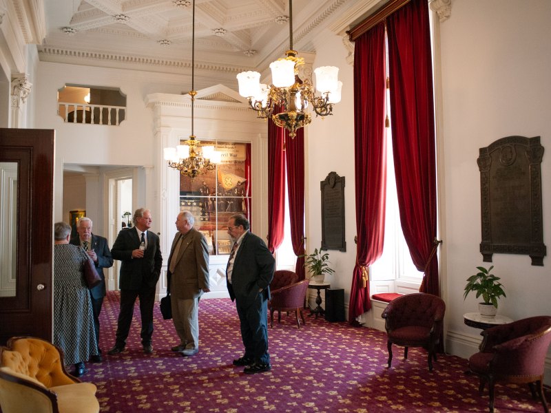 A group of people in formal attire stand and converse in an elegant room with red curtains and ornate chandeliers.