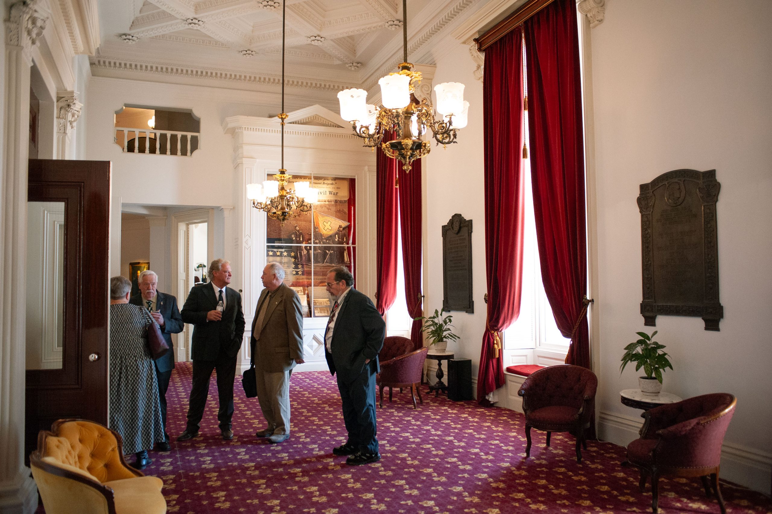 A group of people in formal attire stand and converse in an elegant room with red curtains and ornate chandeliers.