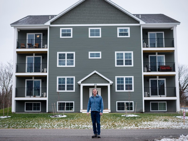 A man stands on a road in front of a four-story green apartment building with balconies, against a cloudy sky.