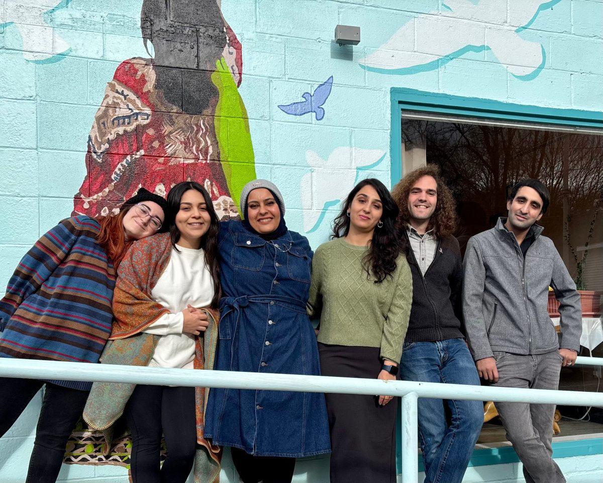 Six people standing together in front of a mural on a light blue wall, smiling at the camera.