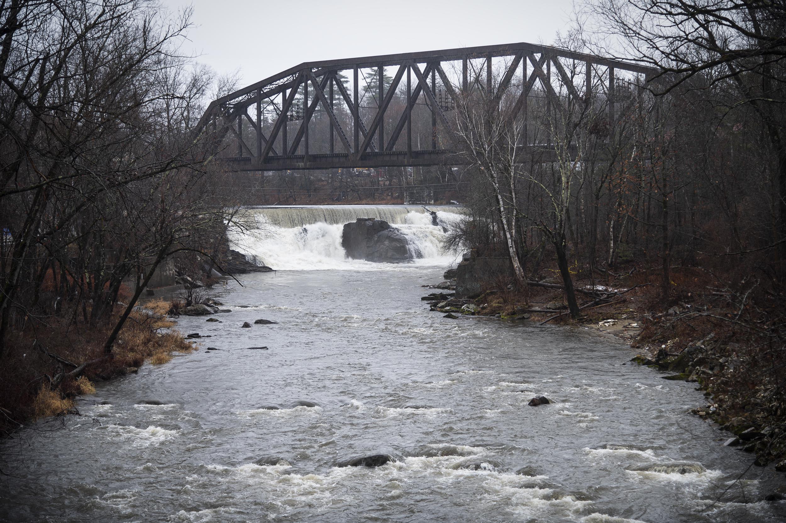 River with a railway bridge above and a waterfall in the background, surrounded by bare trees on an overcast day.