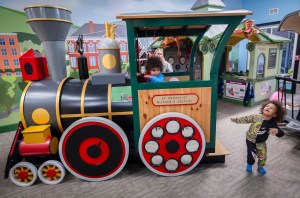 Children play on a colorful, stationary train exhibit inside a play center. A sign on the train reads, "In memory of Richard J. Costello.