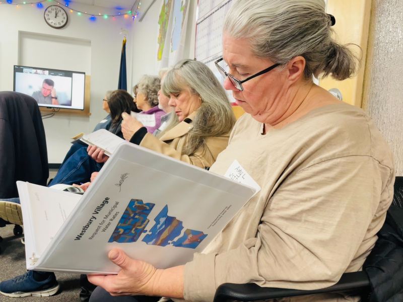 A group of people seated in a room, reading documents titled "Westbury Village," with a video conference screen in the background.