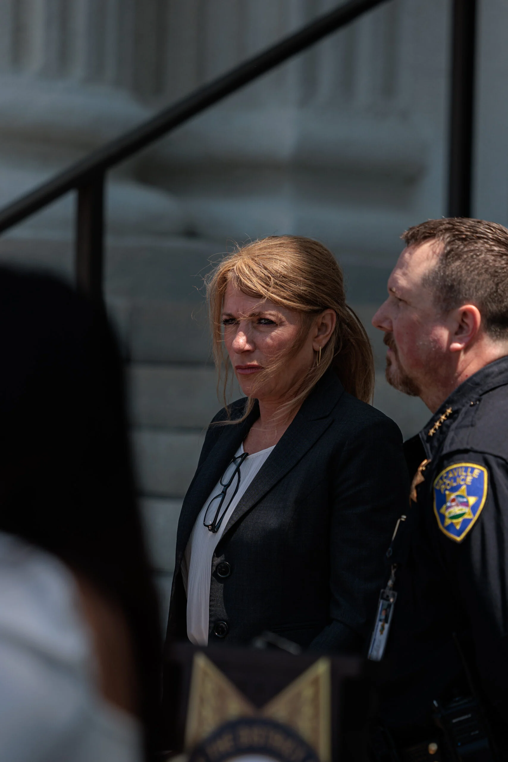 A woman in a dark blazer and glasses stands next to a uniformed police officer in front of a building with columns.