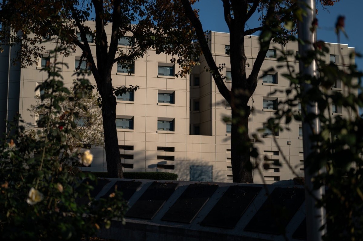A beige building with multiple windows is partially obscured by trees and foliage in the foreground, under a clear blue sky.