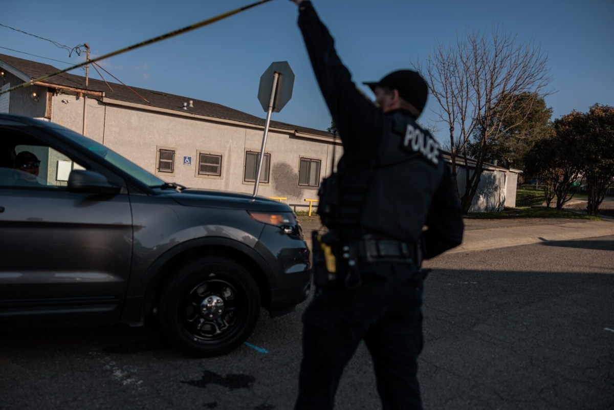 Police officer in uniform holds up tape near a parked SUV on a sunny day, with buildings and a stop sign in the background.