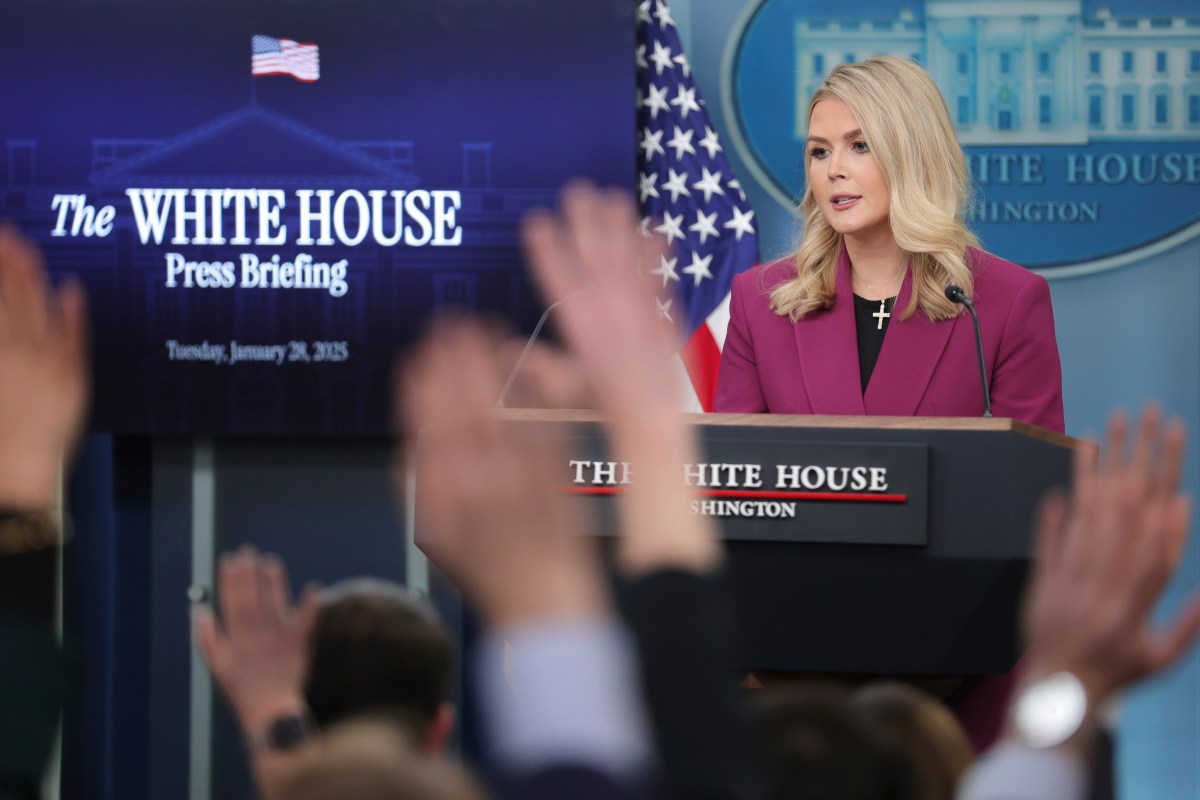 A woman stands at a podium during a White House press briefing. Multiple hands are raised in the foreground.