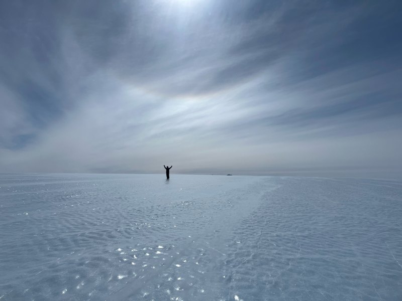 Person stands with arms raised on a vast, reflective icy surface under a cloudy sky, with the sun partially visible.