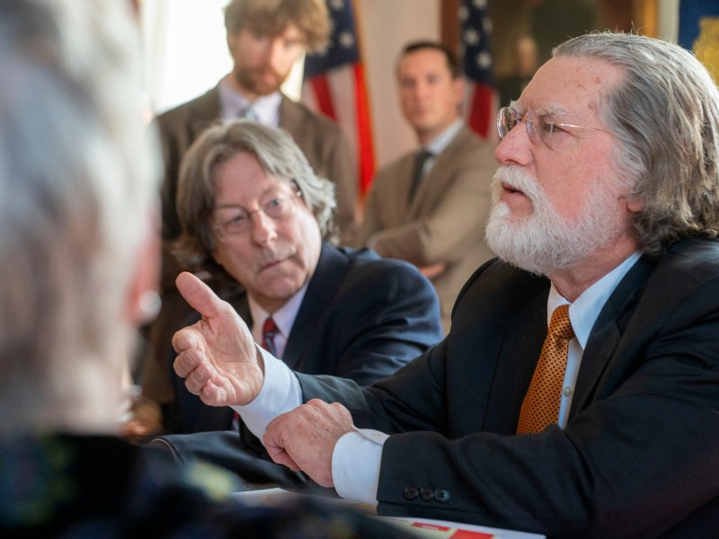 A bearded man in a suit speaks assertively at a meeting, gesturing with his hand. Participants in the background listen. American flags are visible.
