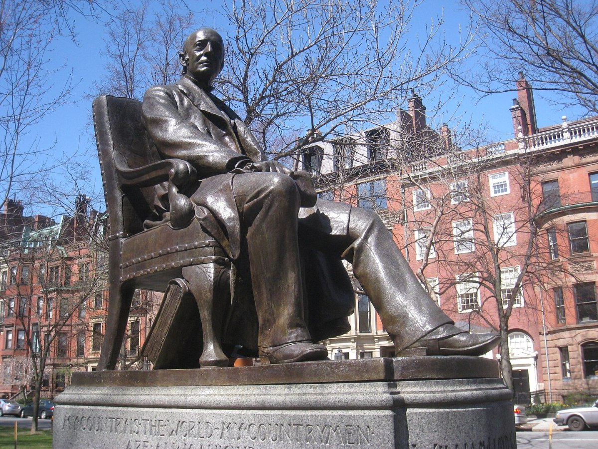 Bronze statue of a seated man in a suit, located on a stone pedestal in an urban park setting with buildings and trees in the background.