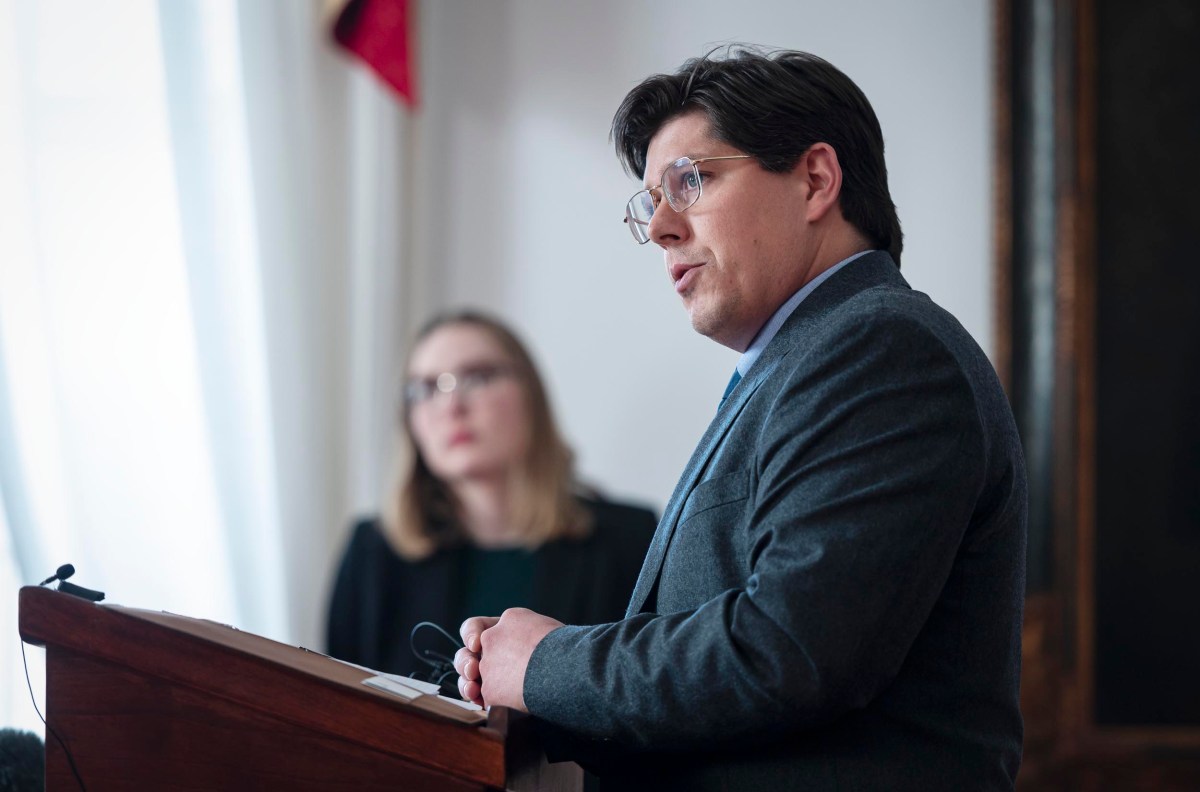 A man in glasses and a suit speaks at a podium. A woman in glasses is blurred in the background.