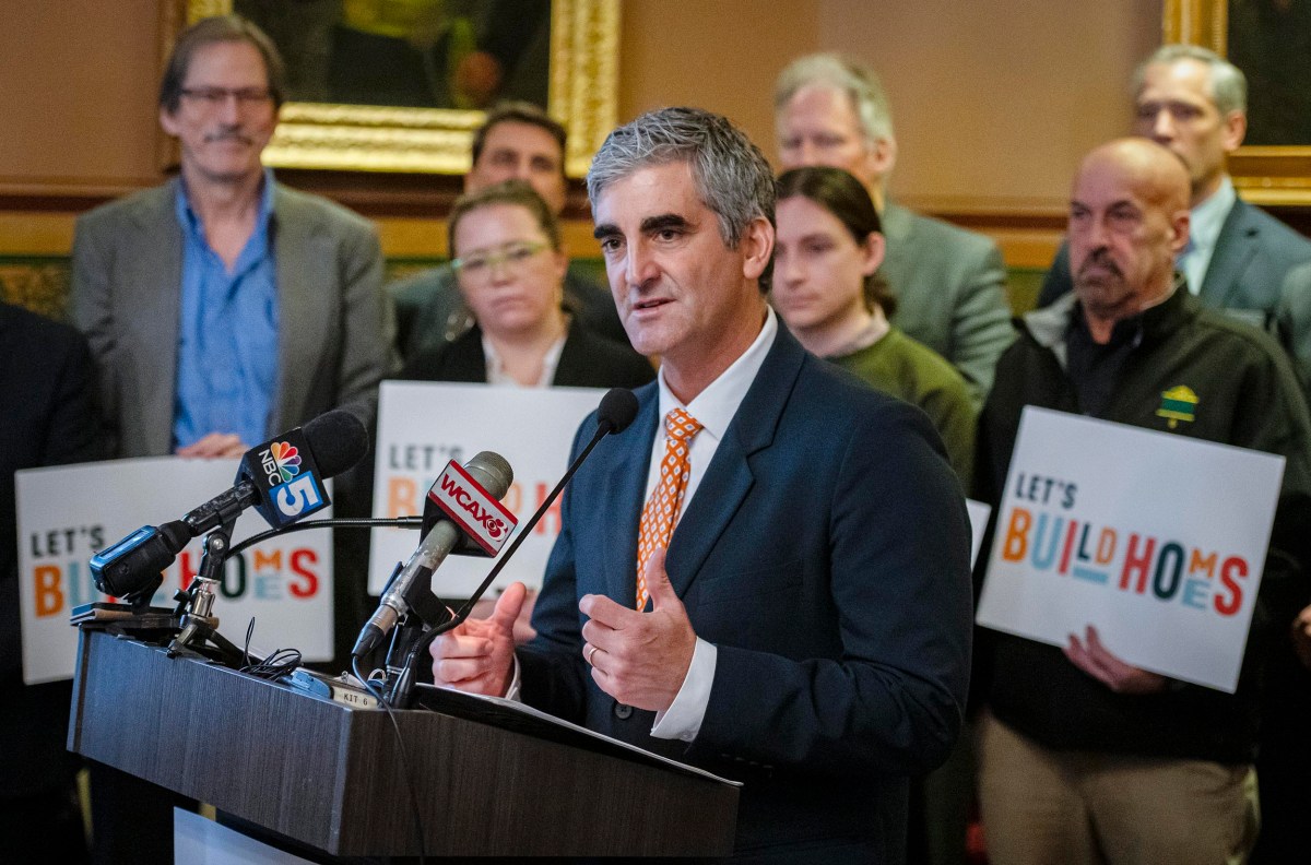 A man speaks at a podium with multiple microphones, surrounded by people holding "Let's Build Homes" signs.