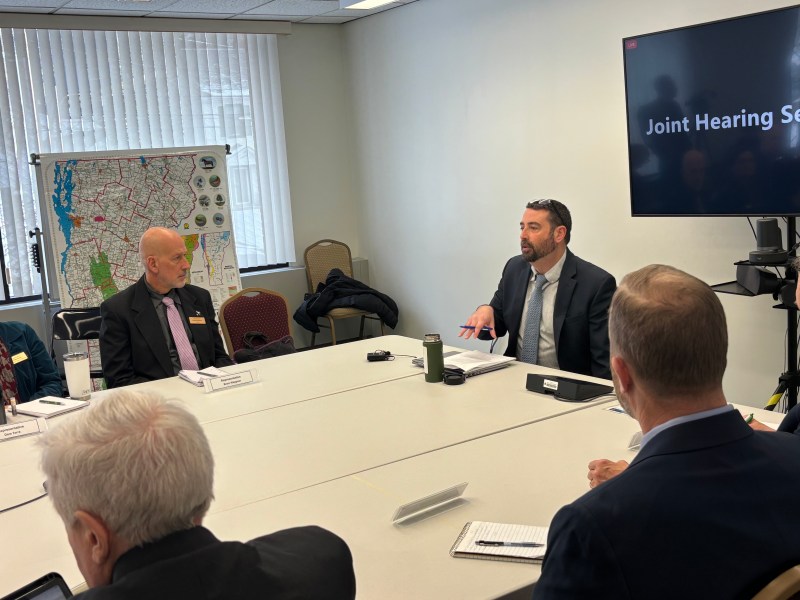 A meeting room with six people seated around a table. A screen displays "Joint Hearing Session." A large map is visible on a stand in the background.