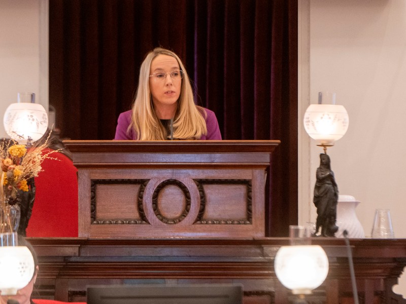 A woman with long hair speaks from a wooden podium in a room with ornate lamps and floral decor.
