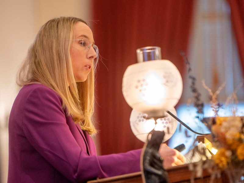 A woman with long blonde hair, wearing glasses and a purple jacket, speaks at a podium. A decorative lamp is in the foreground, with red curtains in the background.