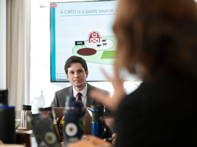 A man in a suit sits at a table with laptops and water bottles. A presentation slide on a screen shows a diagram related to CAFOs (Concentrated Animal Feeding Operations).