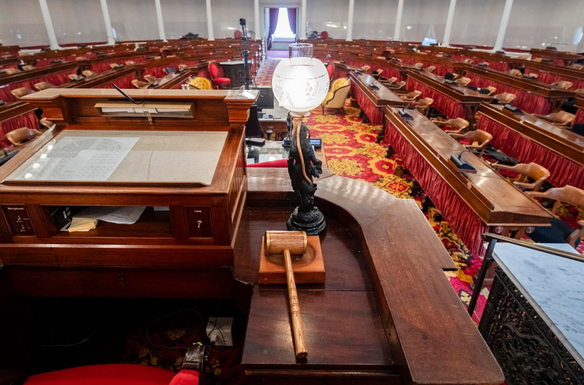 An empty legislative chamber with wooden desks, red chairs, and a gavel on a podium in the foreground.