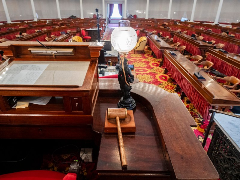 An empty legislative chamber with wooden desks, red chairs, and a gavel on a podium in the foreground.
