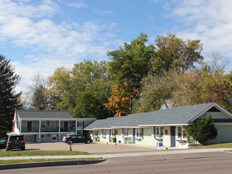 A small roadside motel with two buildings, surrounded by trees under a partly cloudy sky. Three cars are parked in the lot.