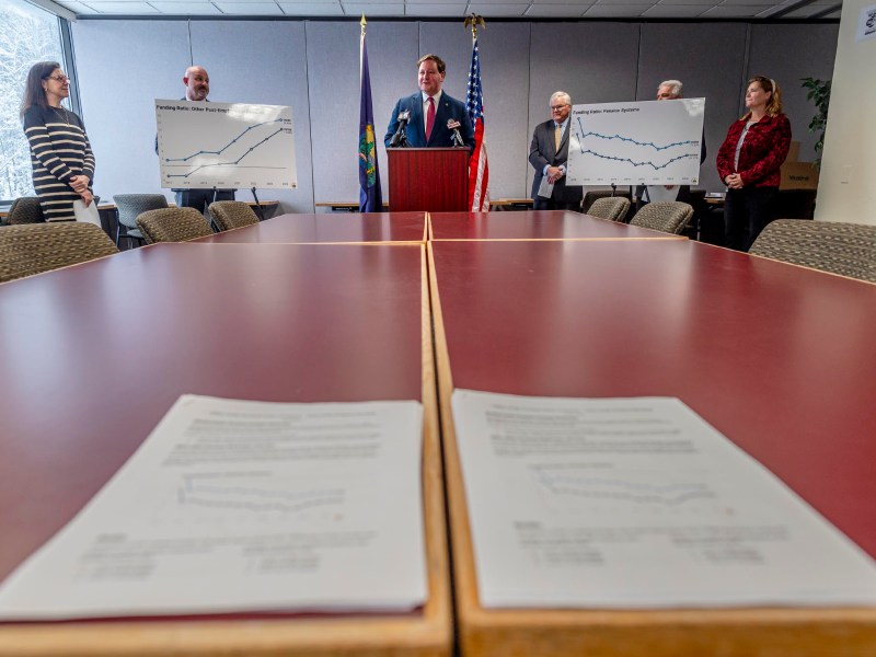 A man speaks at a podium with graphs on easels showing employment trends. Five people stand around him in a conference room. Two documents with graphs lie on a table in the foreground.