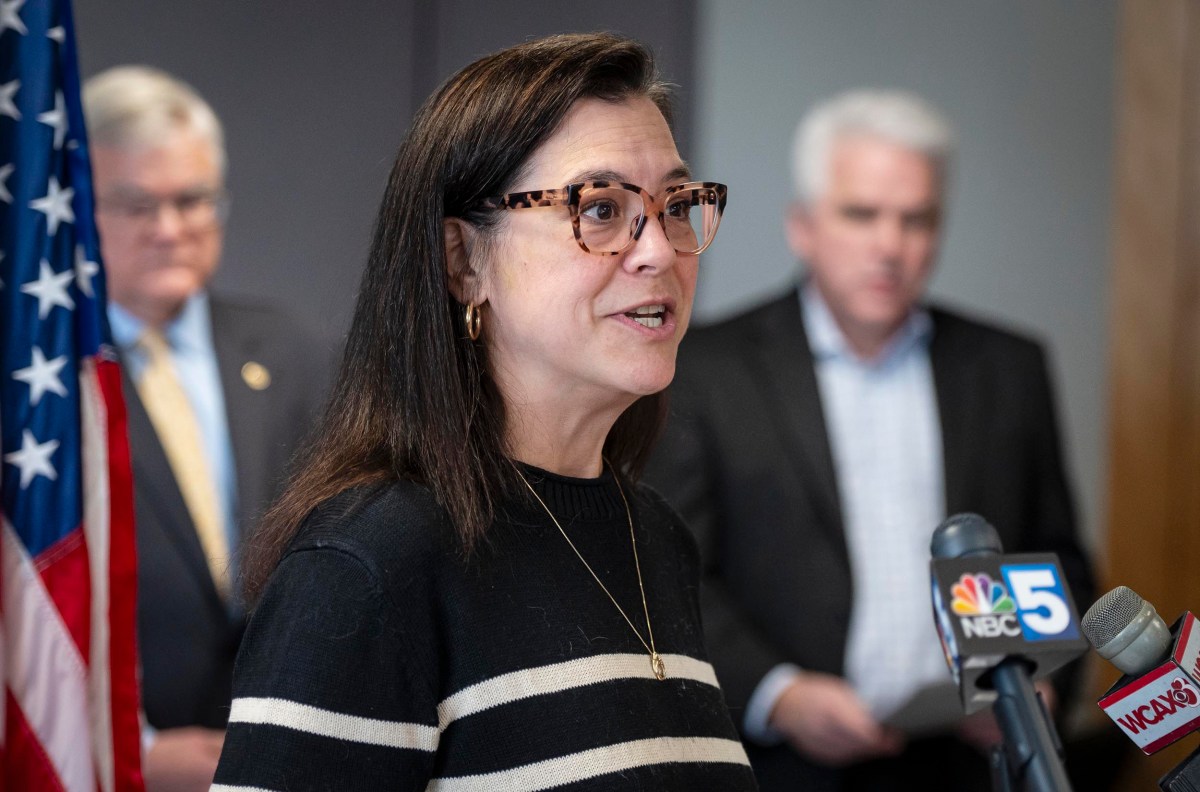A woman in glasses speaks at a podium with microphones from various news outlets. Two men in suits stand in the background near an American flag.