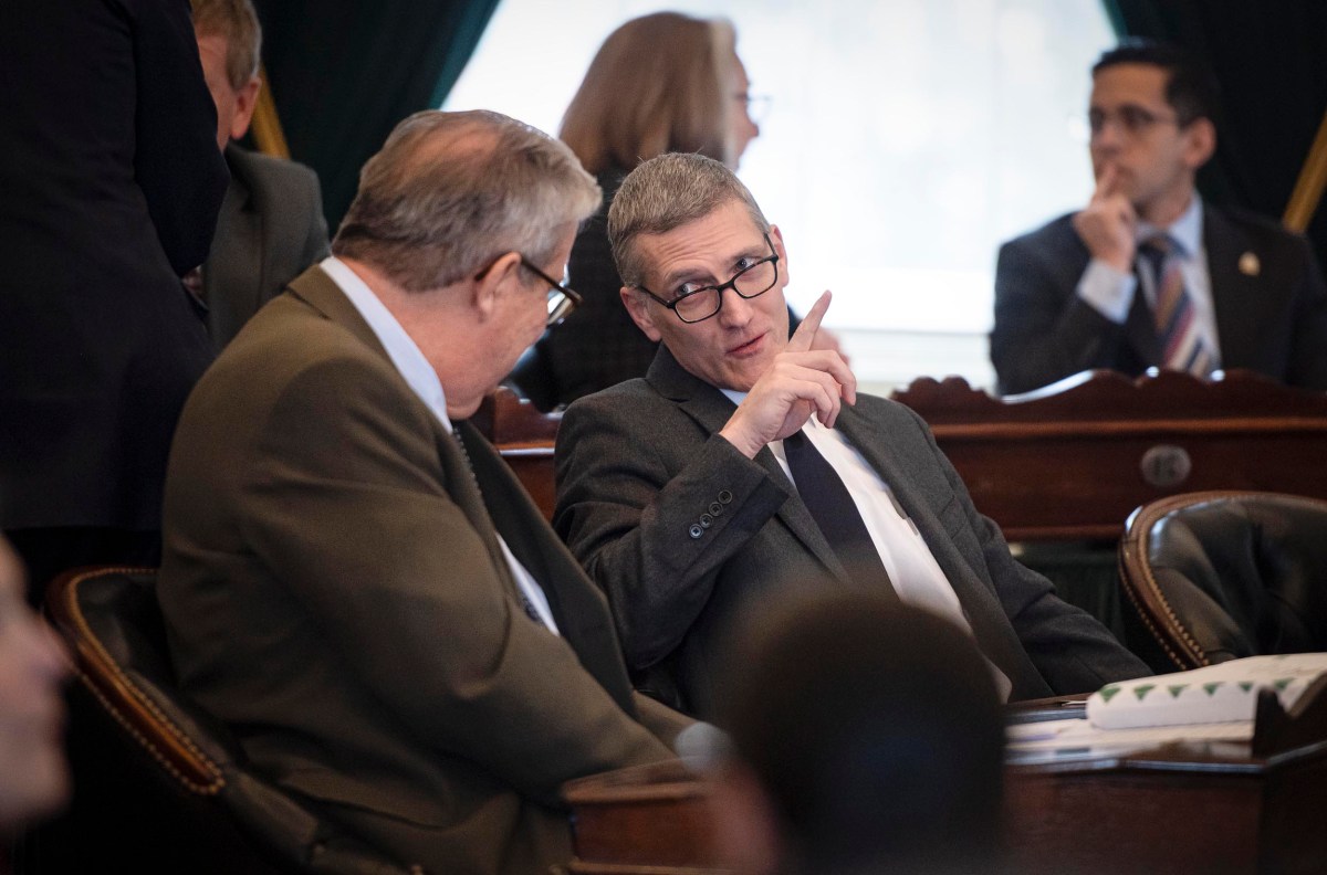 Two men in suits are seated, having a discussion. One gestures with his finger while speaking. Other people are visible in the background.